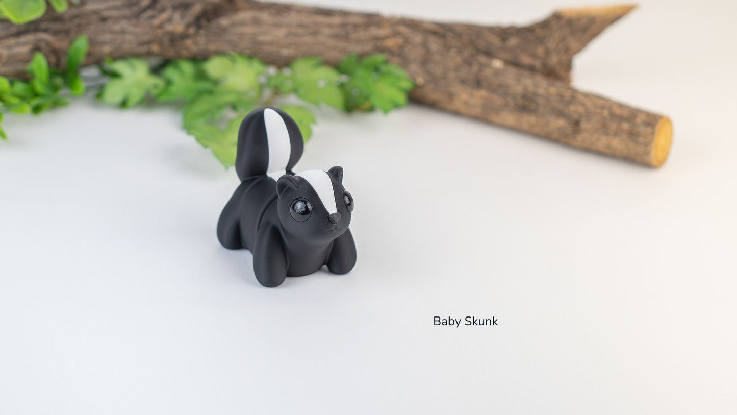 Black baby skunk figurine on a white surface with green leaves and a branch in the background.