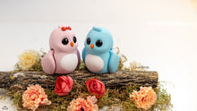 Two small bird figurines, one pink and one blue, sitting on a log with flowers around them.