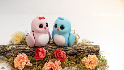 Two small bird figurines, one pink and one blue, sitting on a log with flowers around them.