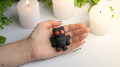 Hand holding a small black creature figurine with red eyes against a white background with candles and greenery.
